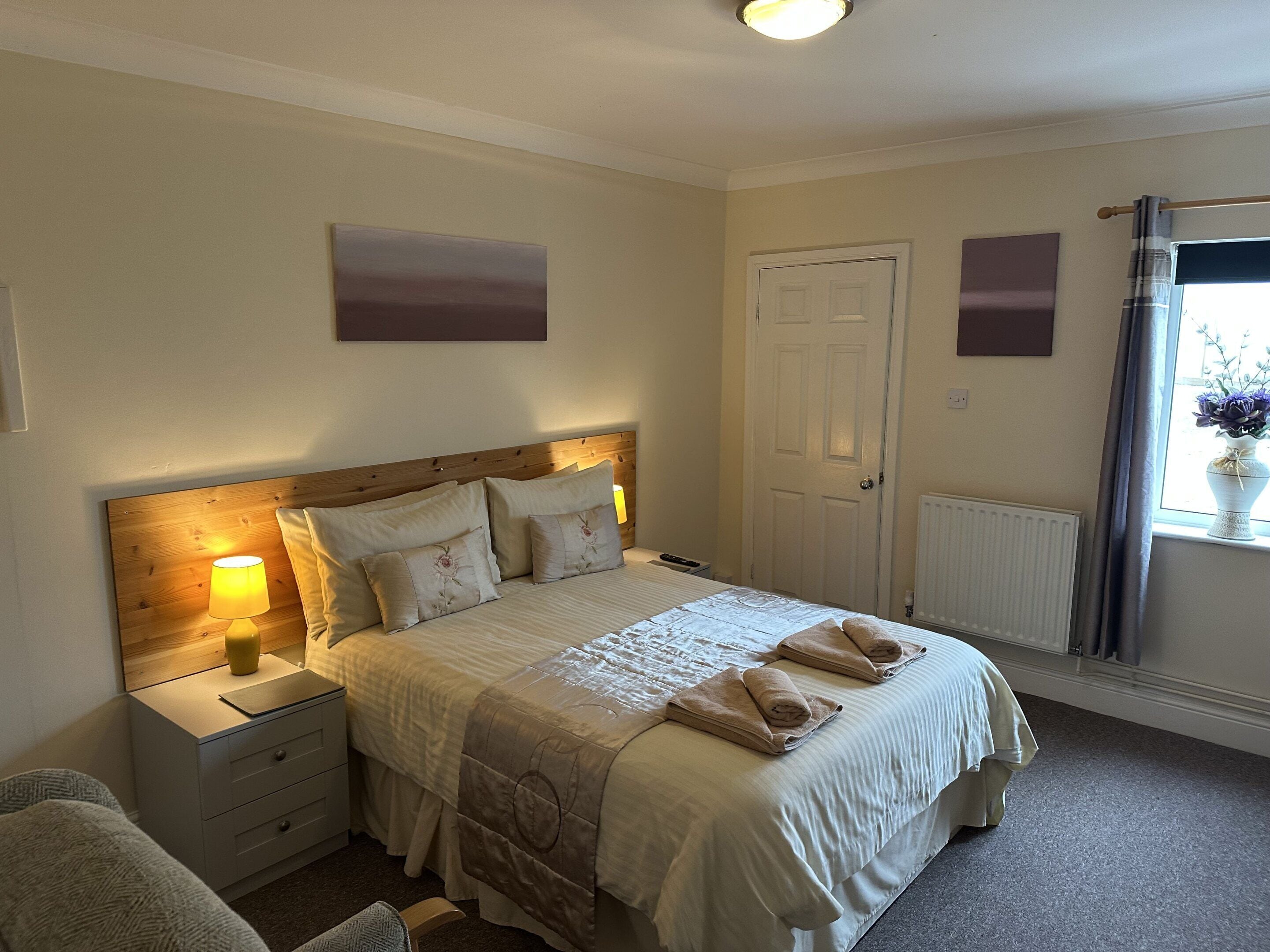 A neatly arranged bedroom with a bed, bedside tables, a nightstand, a chair, a door, and a window with curtains.