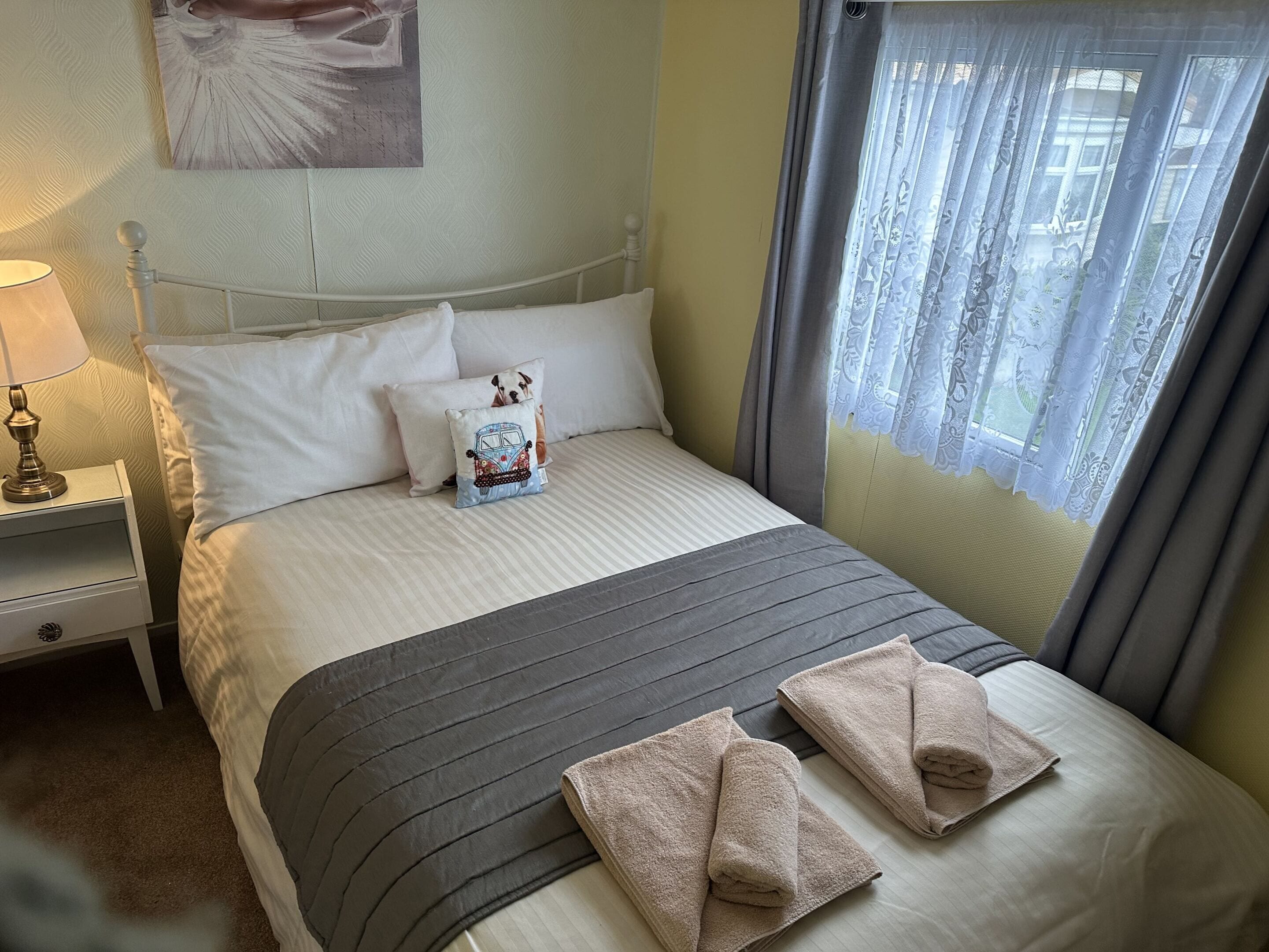 Een netjes opgemaakt bed met een gestreept dekbed, kussens en een sierkussen. Er is een nachtkastje met een lamp en een lade. De kamer heeft een raam met gordijnen.