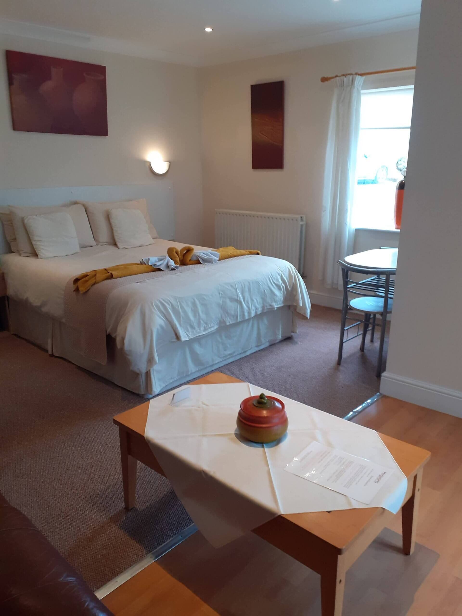 A hotel room with a bed, a table with a red object, a chair, and a window with curtains.