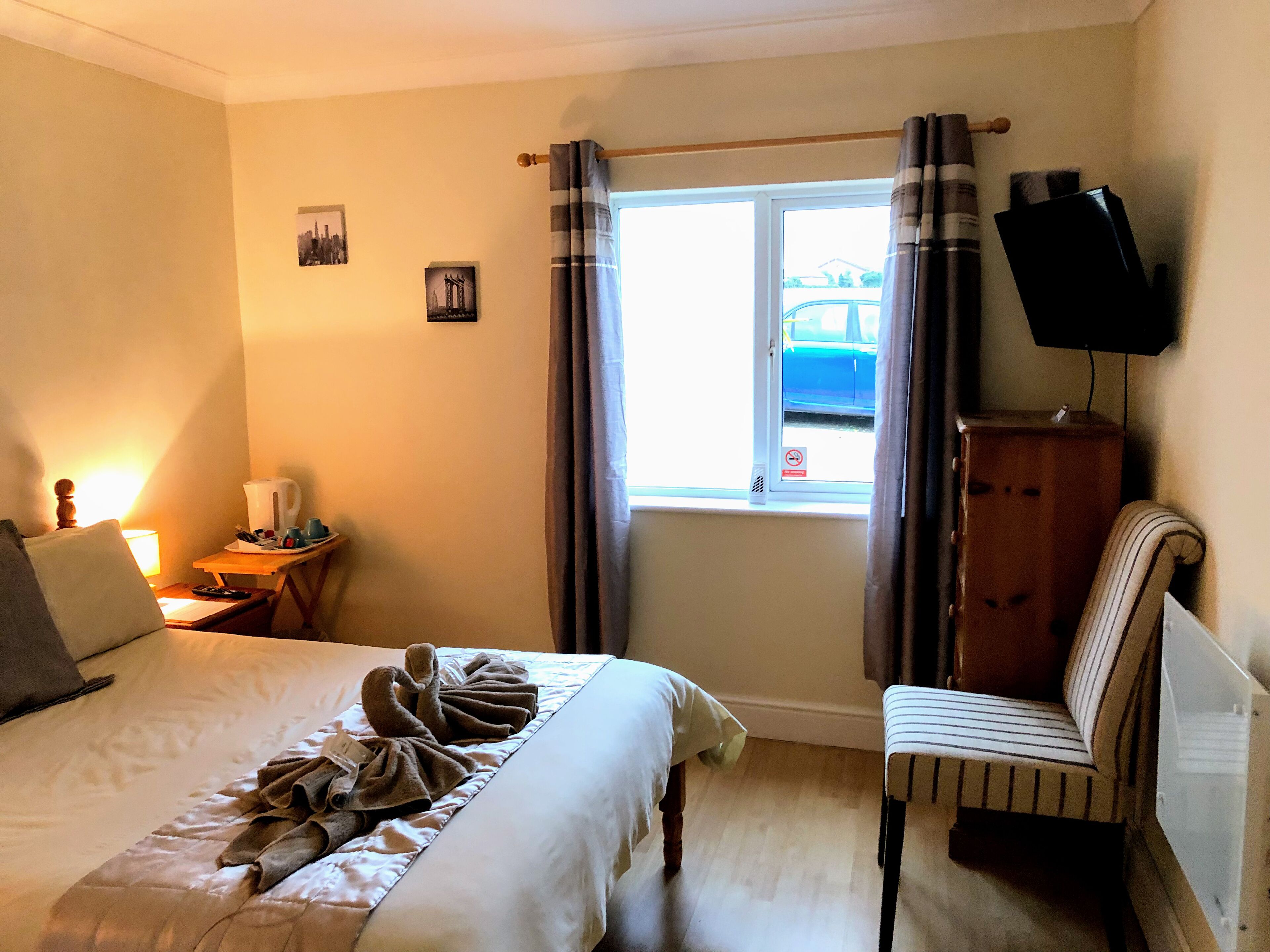 A hotel room with a bed, a chair, a TV, a window with curtains, and a small table with a lamp and a kettle.