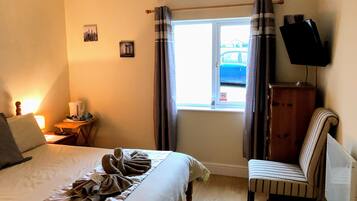 A hotel room with a bed, a chair, a TV, a window with curtains, and a small table with a lamp and a kettle.