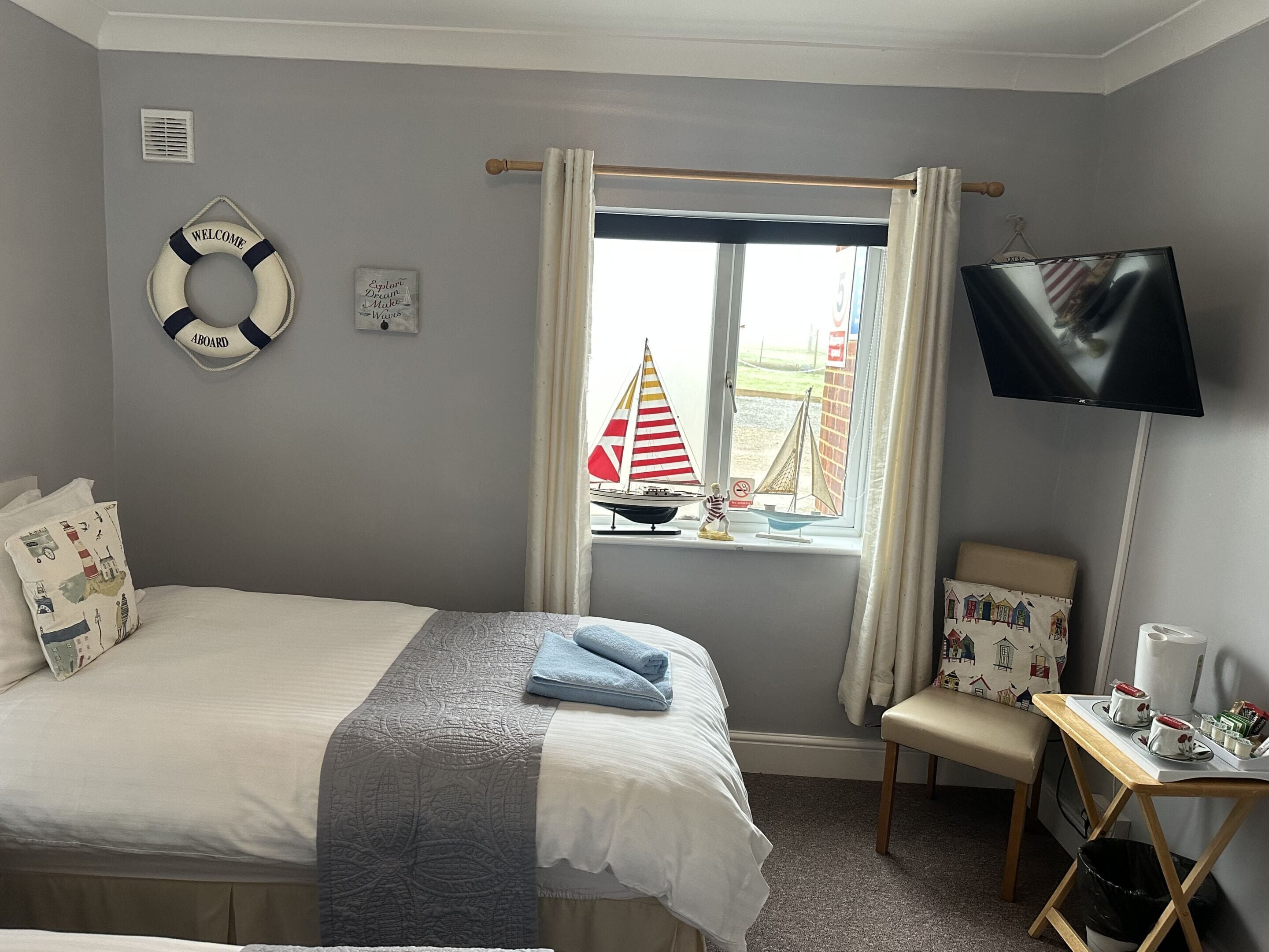 A bedroom with a bed, a small table with a TV, a window with curtains, a life ring, and a sign saying "Explore Dream Make Waves".