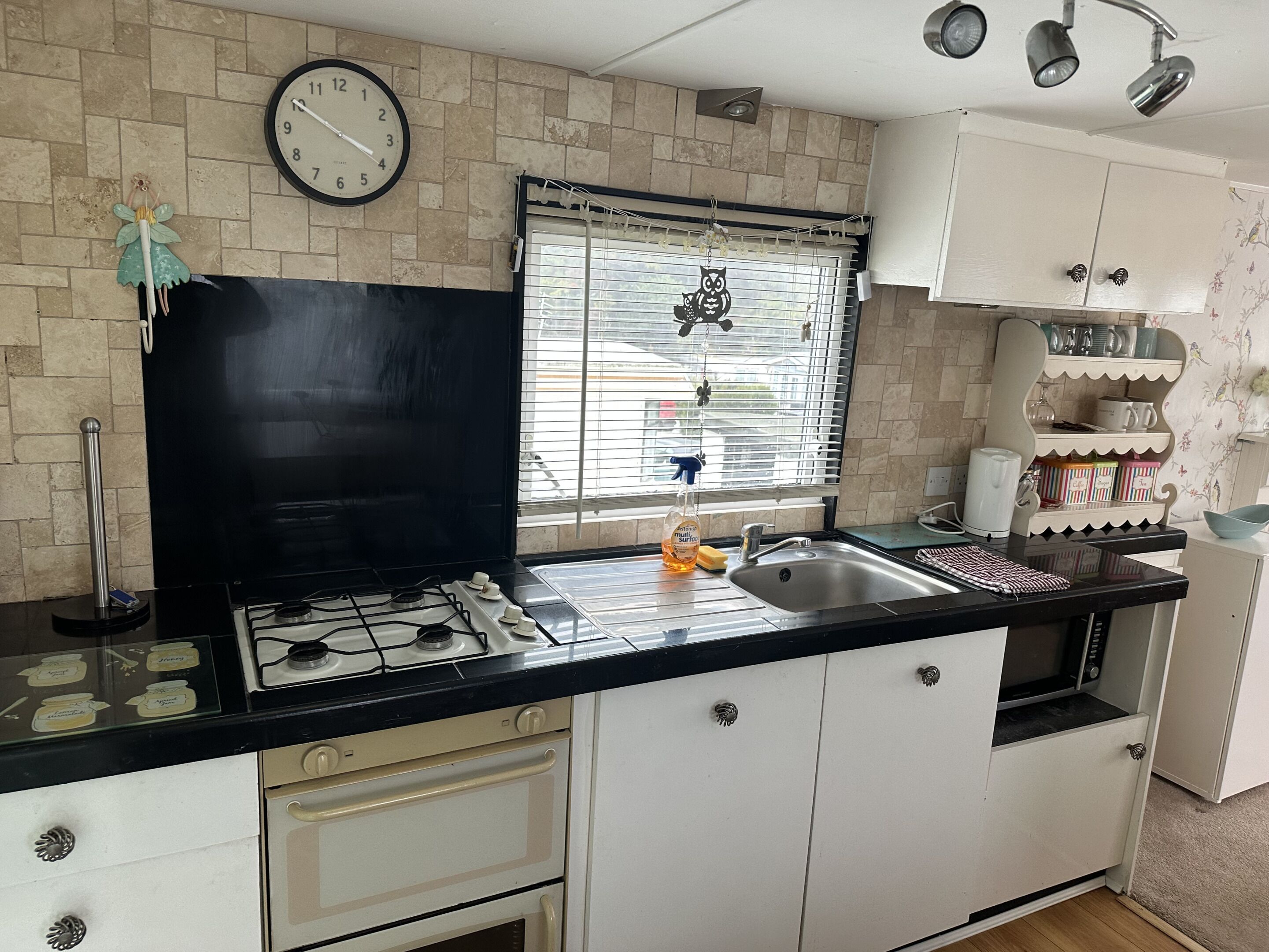 Cocina con estufa a gas, lavamanos y armarios blancos. Hay un reloj en la pared y una ventana con persianas.