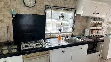 Cocina con estufa a gas, lavamanos y armarios blancos. Hay un reloj en la pared y una ventana con persianas.
