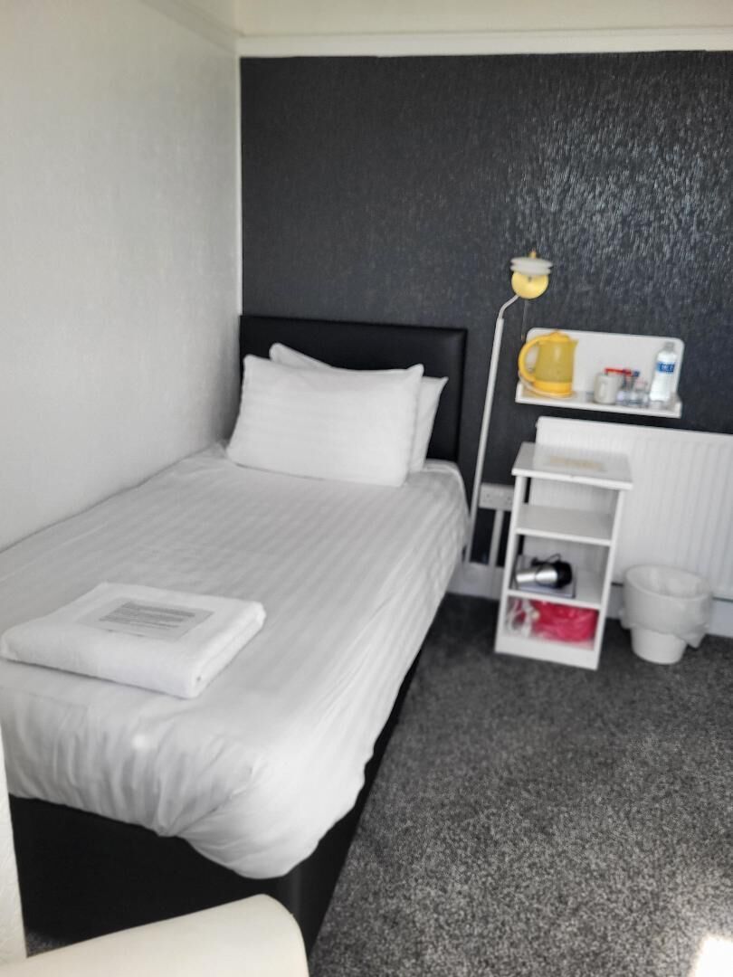 A hotel room with a bed, a nightstand, a lamp, a shelf with toiletries, and a radiator.