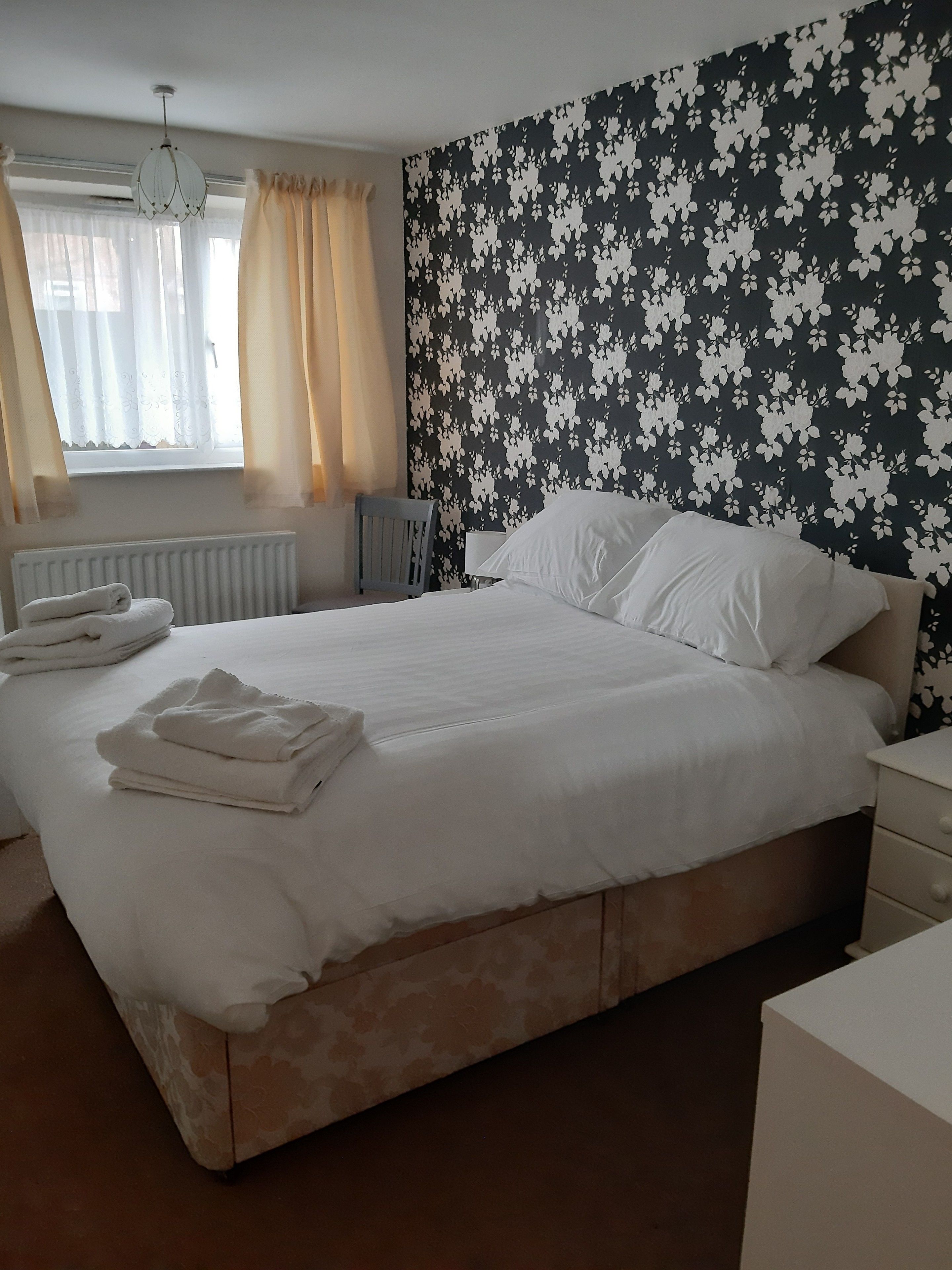 A neatly made bed with white linens and folded towels, a bedside table, and a wall with a floral pattern.