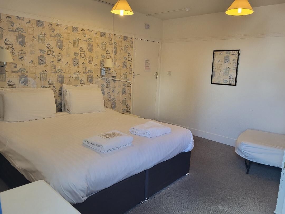 A neatly made bed with white linens and towels, a bedside table, a wall with a bird pattern wallpaper, and a framed picture on the wall.