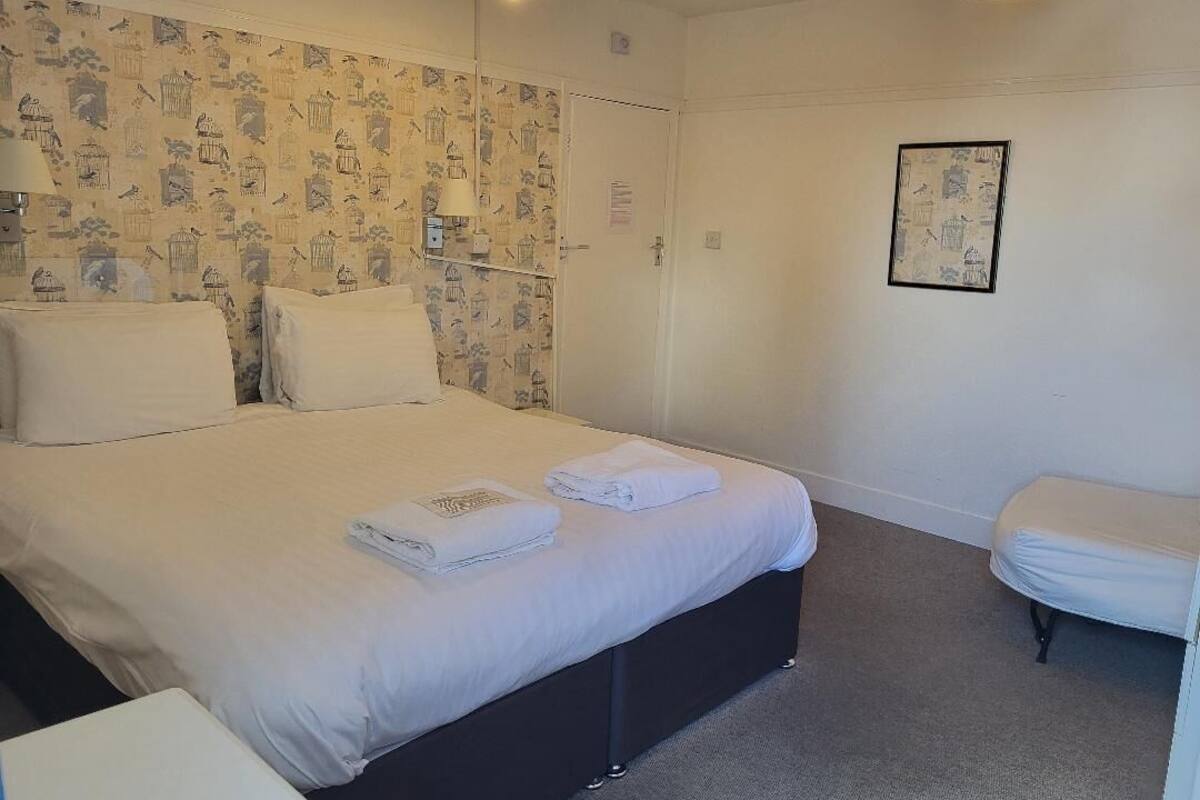 A neatly made bed with white linens and towels, a bedside table, a wall with a bird pattern wallpaper, and a framed picture on the wall.