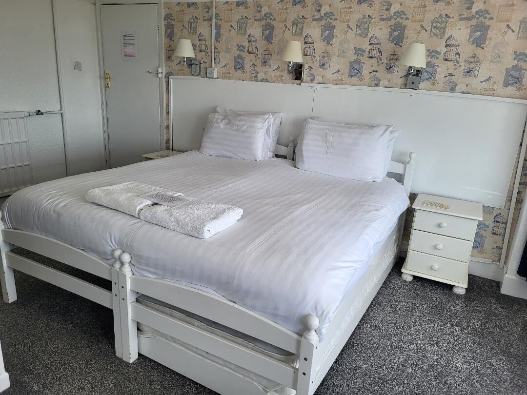 A neatly made bed with white linens and pillows, a bedside table with a lamp, and a wall with a bird pattern wallpaper.