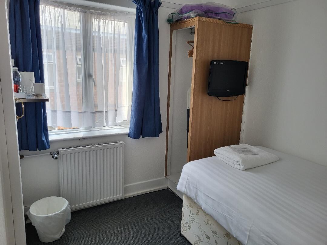 A small, tidy room with a bed, a wooden wardrobe, a television mounted on the wall, a radiator, a window with curtains, and a small trash bin.