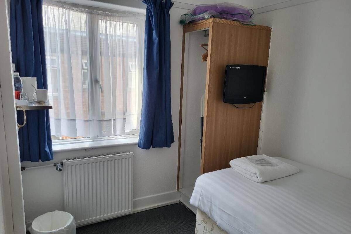 A small, tidy room with a bed, a wooden wardrobe, a television mounted on the wall, a radiator, a window with curtains, and a small trash bin.