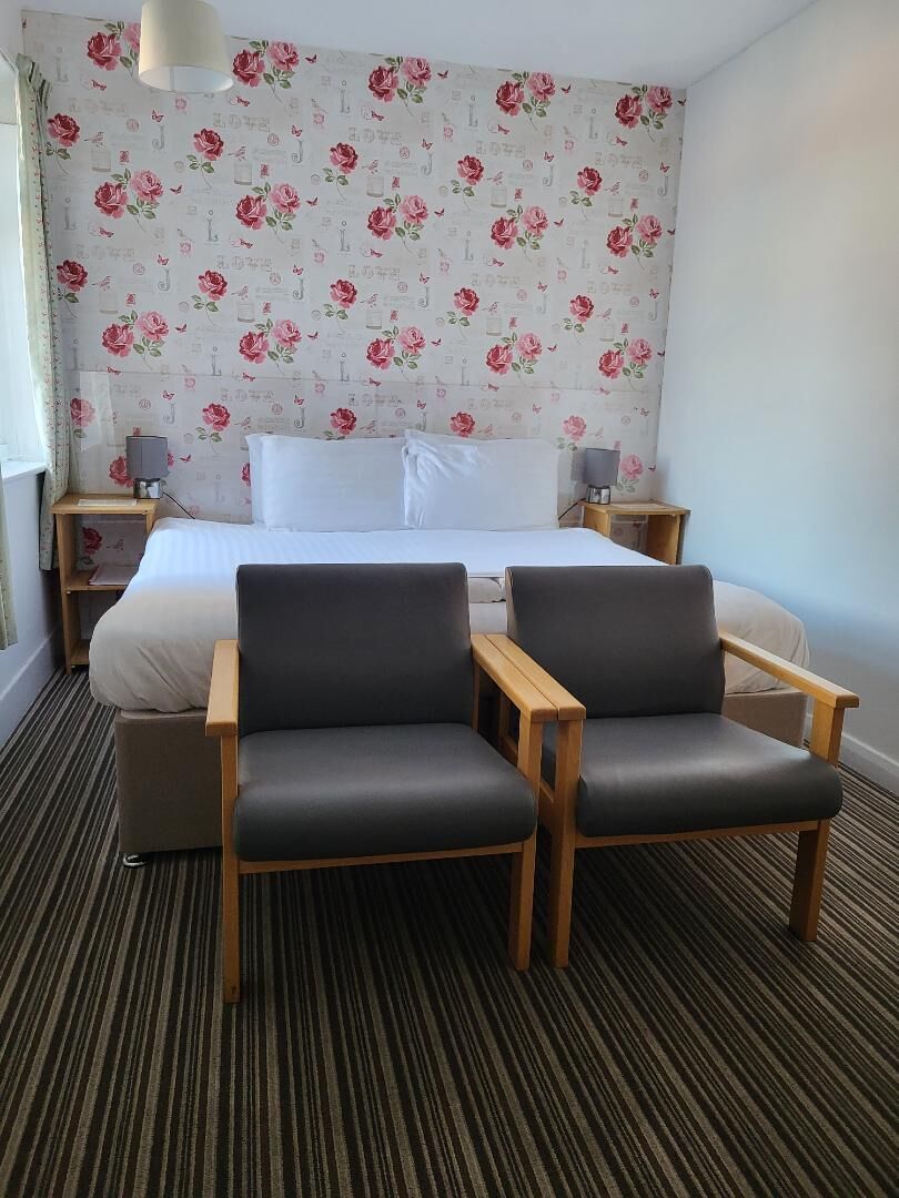 A hotel room with a bed, two armchairs, a nightstand, a lamp, and a floral wallpaper.