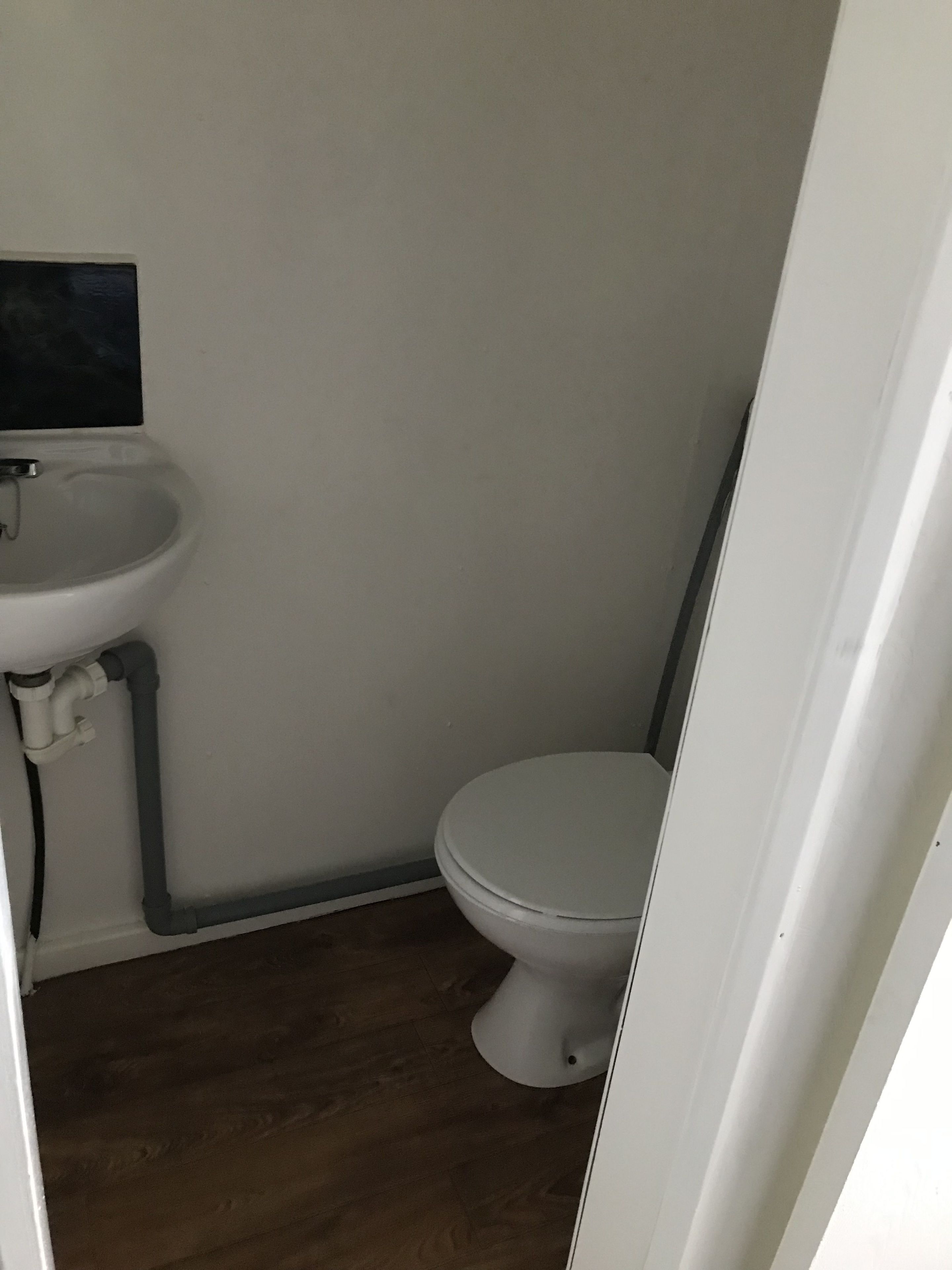 Uma casa de banho pequena e compacta, com uma sanita e lavatório brancos, soalho em madeira e uma parede simples.