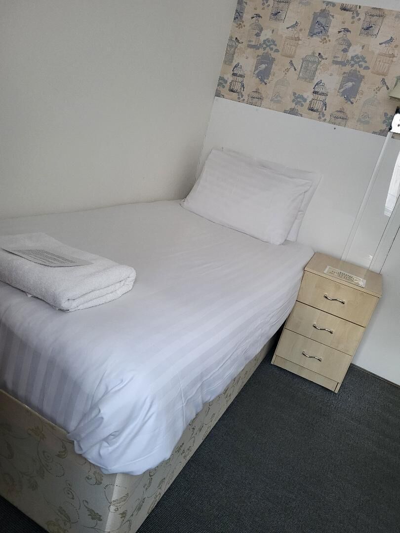 Ein Einzelbett mit weißer Bettdecke und einem gefalteten Handtuch, ein Nachttisch aus Holz mit Schubladen und eine Wand mit Vogelkäfig-Muster.