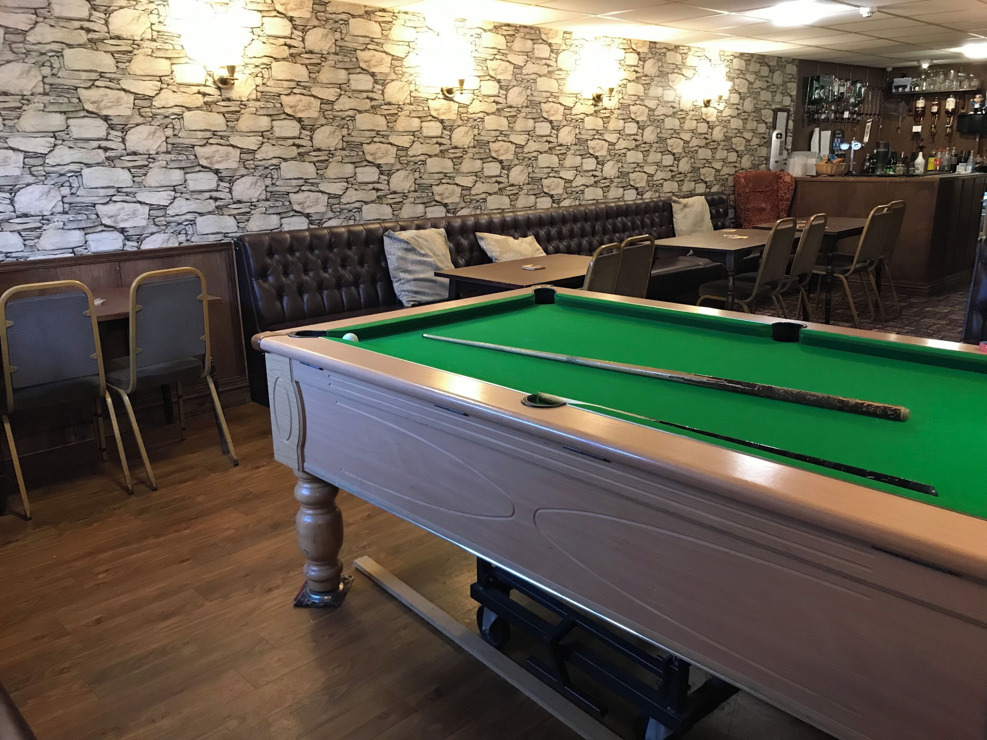 A billiards table with a green felt surface, a pool cue, and a cue ball. There is a stone wall with a textured pattern and wall-mounted lighting fixtures.