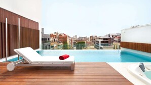 A rooftop pool area with a lounge chair and a red towel, wooden deck, and a view of the cityscape.