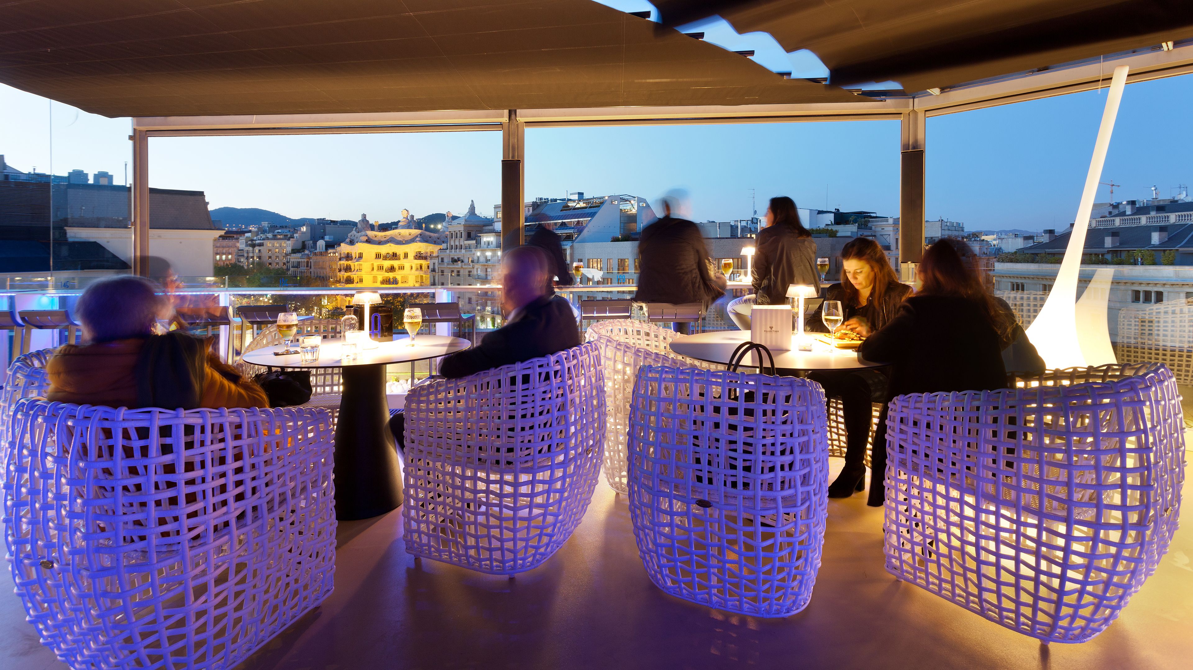 A rooftop terrace with wicker chairs and tables, overlooking a cityscape.