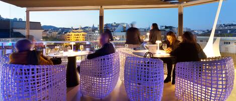 A rooftop terrace with wicker chairs and tables, overlooking a cityscape.