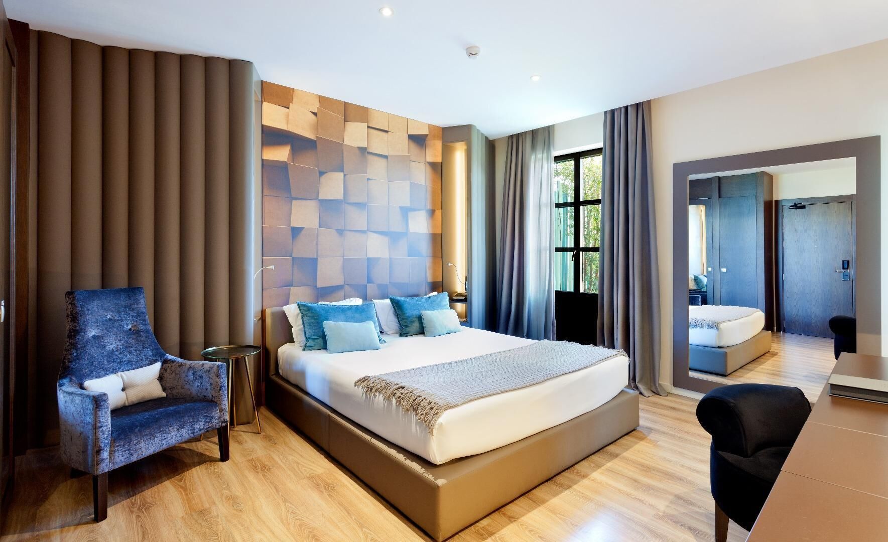 A modern hotel room with a large bed, a seating area with a chair, a small table, and a mirror.