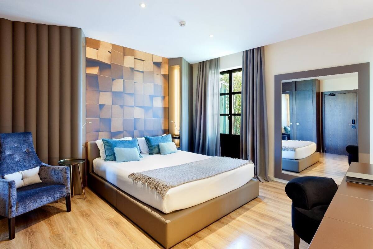 A modern hotel room with a large bed, a seating area with a chair, a small table, and a mirror.