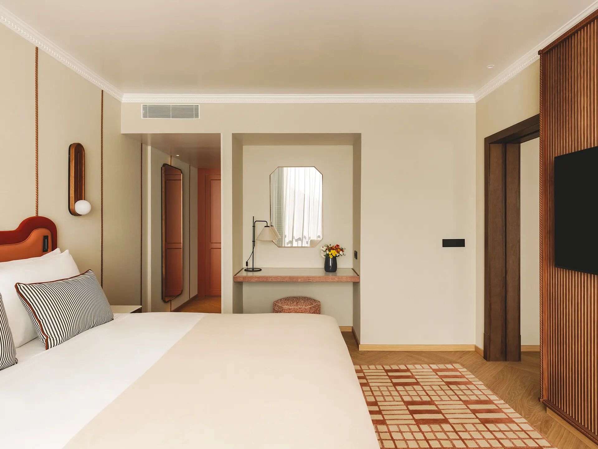 Una habitación de hotel con cama, una mesita de noche con lámpara, espejo, un pequeño jarrón con flores y televisor.