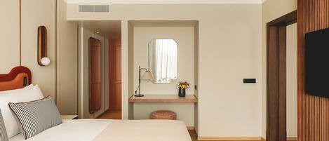 Una habitación de hotel con cama, una mesita de noche con lámpara, espejo, un pequeño jarrón con flores y televisor.