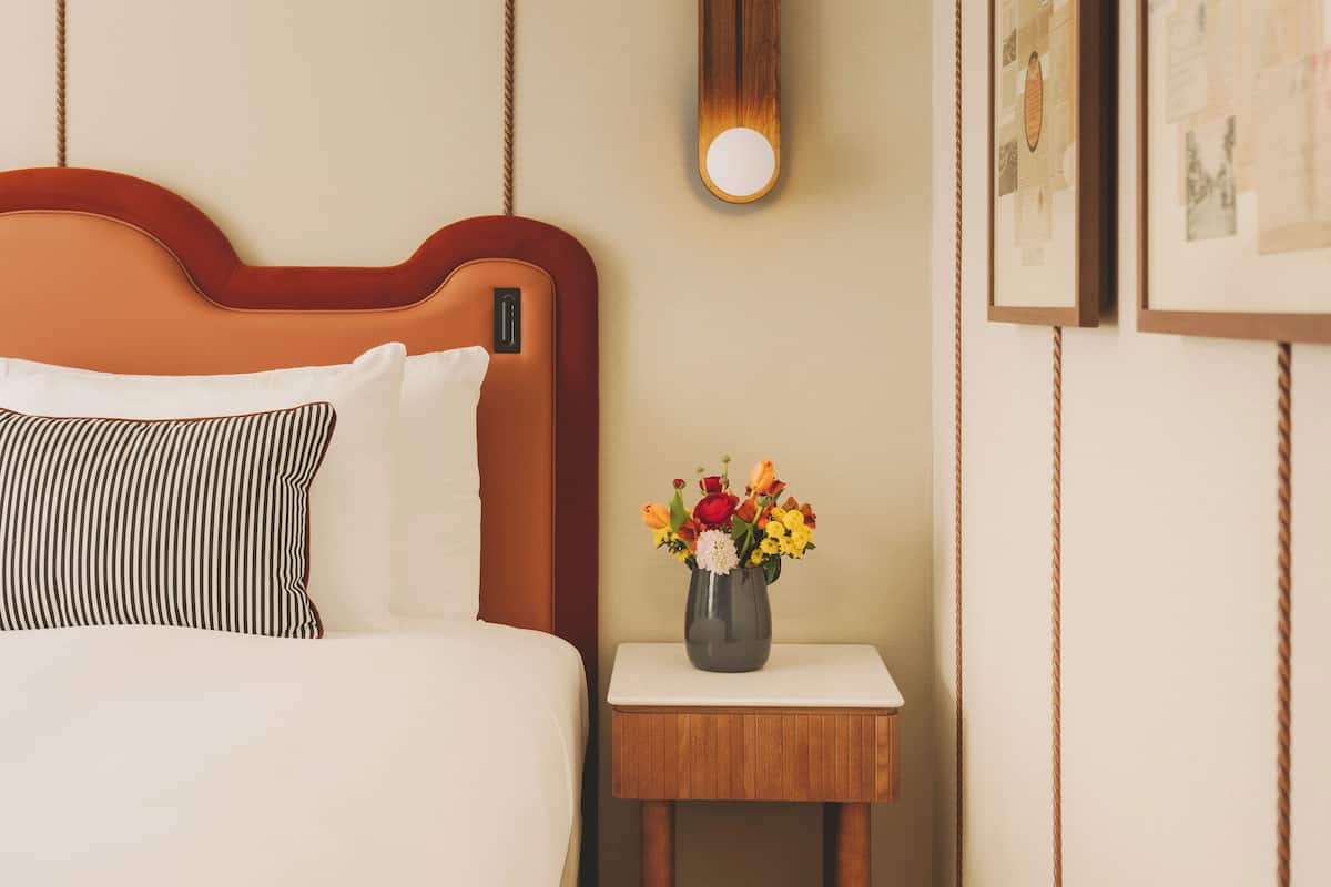 Una habitación de hotel con una cama, una mesita de noche con un florero y un reloj de pared, además de cuadros enmarcados.