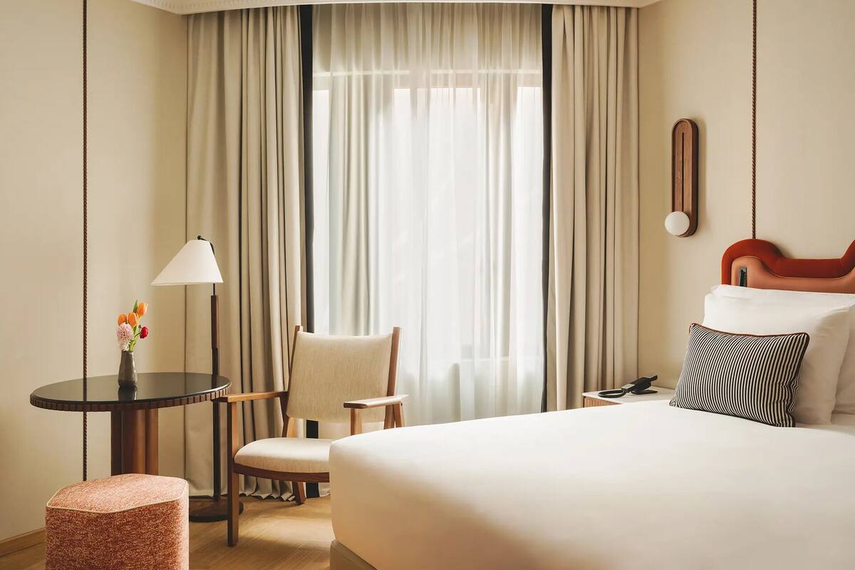 Una habitación de hotel con una cama, una silla, una mesita con lámpara y una ventana con cortinas.