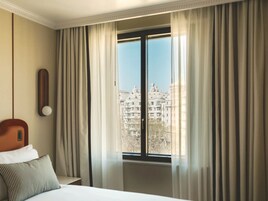 Una habitación de hotel con cama, mesita de noche, lámpara y ventana con cortinas.