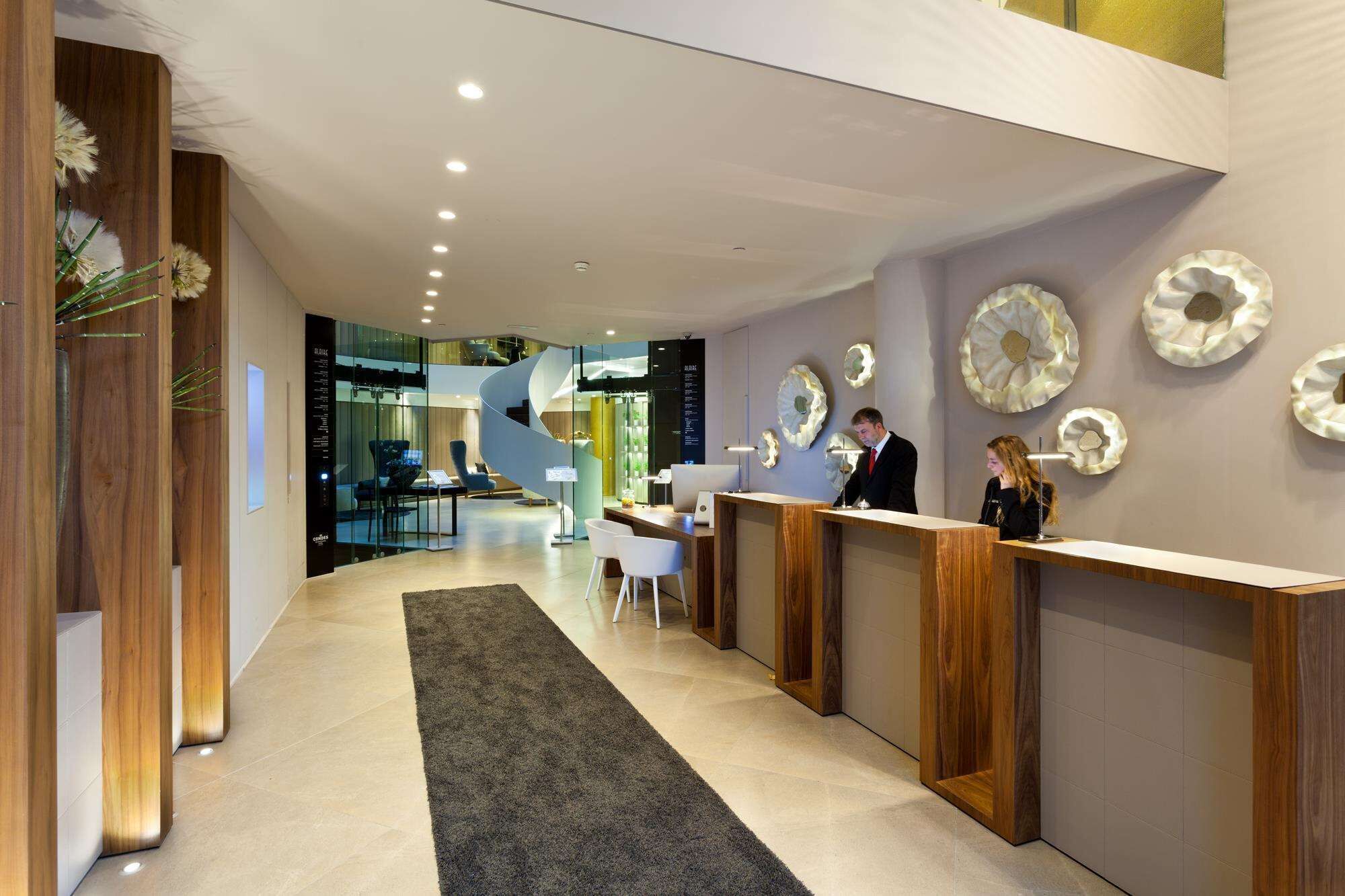Een moderne hotelontvangsthal met een centrale trap, houten receptiebalies en decoratieve wandkunst.
