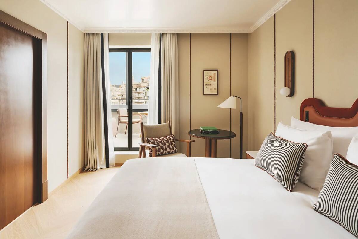 Habitación de hotel con una cama grande, una mesita pequeña, una silla, un balcón con vistas y un cuadro en la pared.