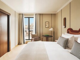 Quarto de hotel com uma cama grande, uma mesa pequena, uma cadeira, varanda com vista e uma moldura na parede.