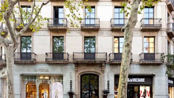 Un edificio de varios pisos con fachada clásica, destacándose una entrada imponente con un gran arco y locales comerciales, incluyendo una tienda de moda de lujo y un hotel.