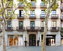 A multi-story building with a classical facade, featuring a prominent entrance with a large arched doorway, and storefronts including a high-end fashion store and a hotel.