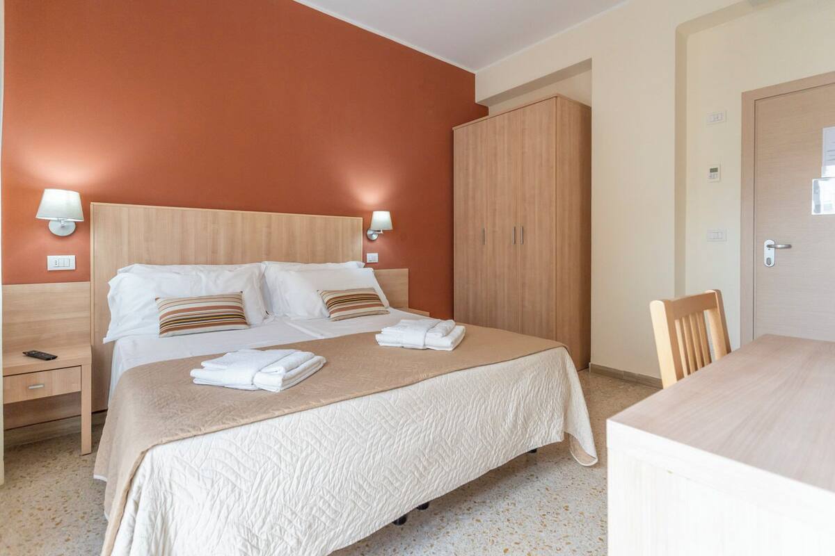 Quarto de hotel com uma cama grande, duas mesas de cabeceira, um guarda-roupa e uma escrivaninha com uma cadeira.