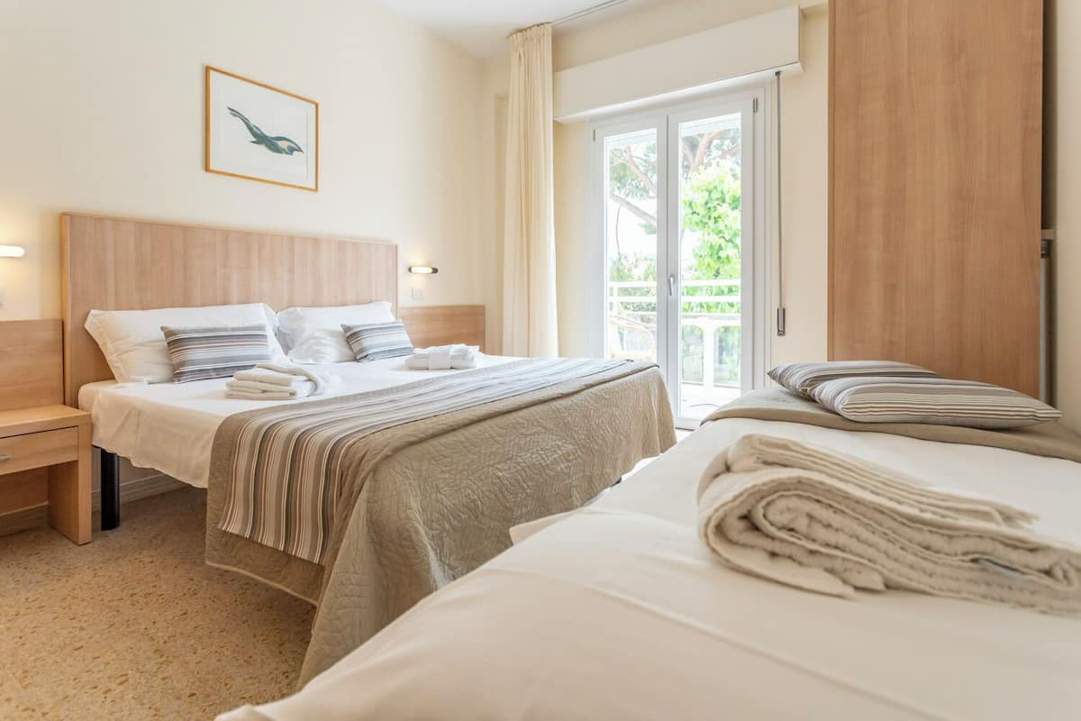 Quarto de hotel com duas camas, cabeceira de madeira, um quadro na parede e vista para área verde pela janela.