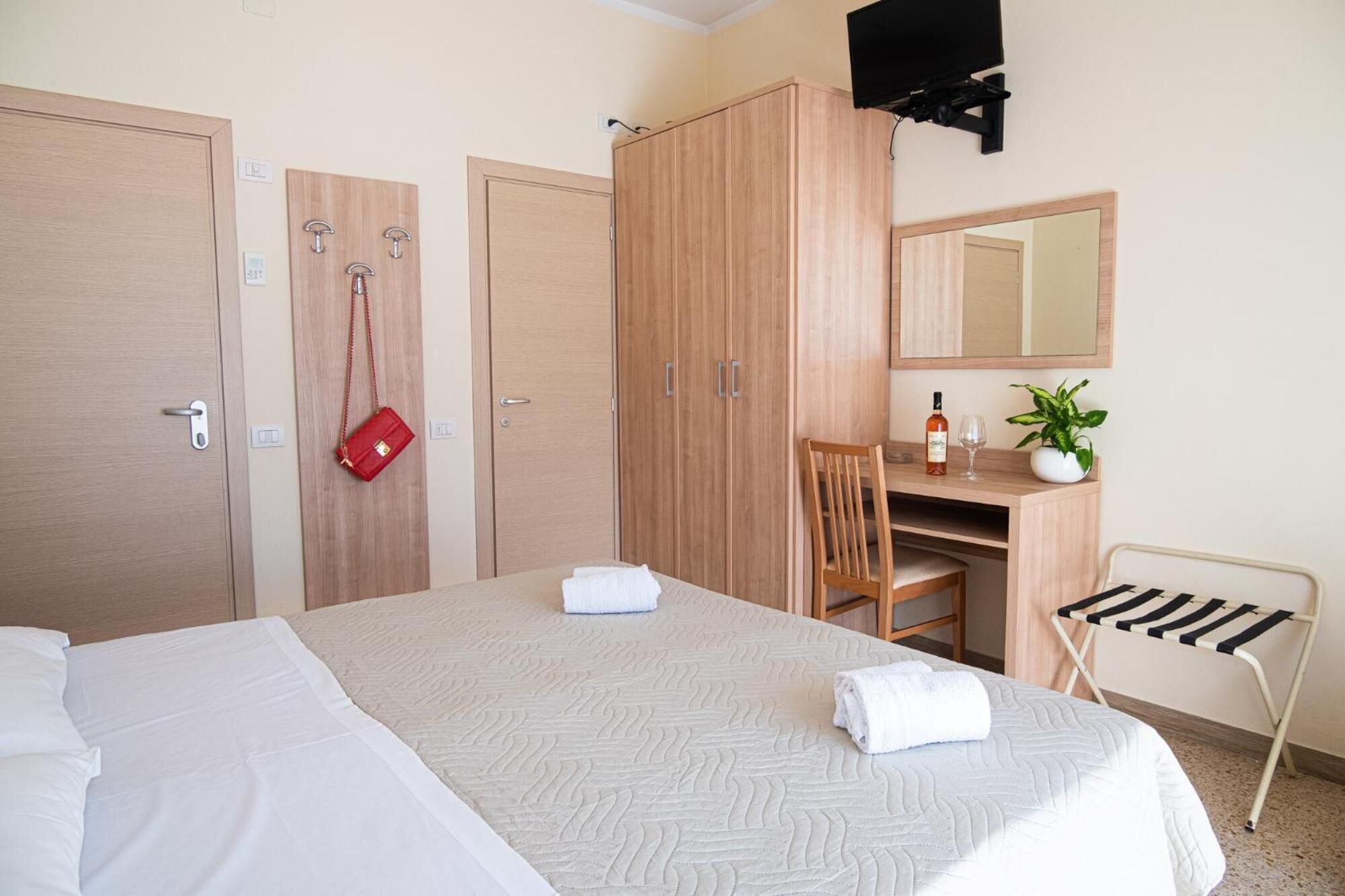 A hotel room with a bed, a desk, a chair, a small table with a bottle and glass, a red bag hanging on the wall, and a TV mounted on the wall.