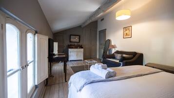 Habitación de hotel con una cama grande, un escritorio, una silla y una mesita. Ventanas en un lateral y una puerta que da al baño.