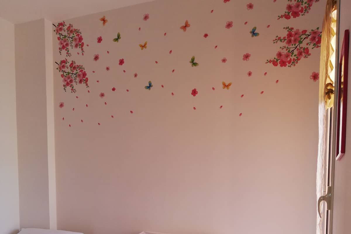 Habitación de hotel con dos camas, una mesita de noche con lámpara, una pared decorativa con diseños florales y de mariposas, y una planta en maceta con flores moradas.