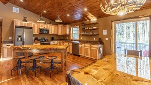 A spacious kitchen with wooden cabinets, a central island with stools, a stainless steel refrigerator, and a large window overlooking a forest. - The Beautiful Whitetail Ridge-Luxury 5BR Cabin, Just minutes from downtown (Wisconsin Dells)