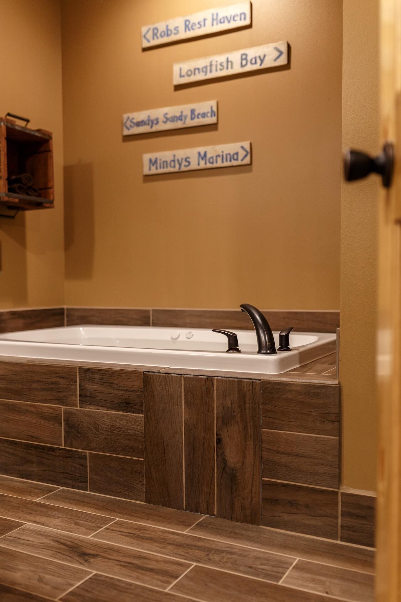 A bathroom with a wooden floor, a bathtub, and directional signs for 'Robs Rest Haven', 'Longfish Bay', 'Sandy's Sandy Beach', and 'Mindys Marina'.