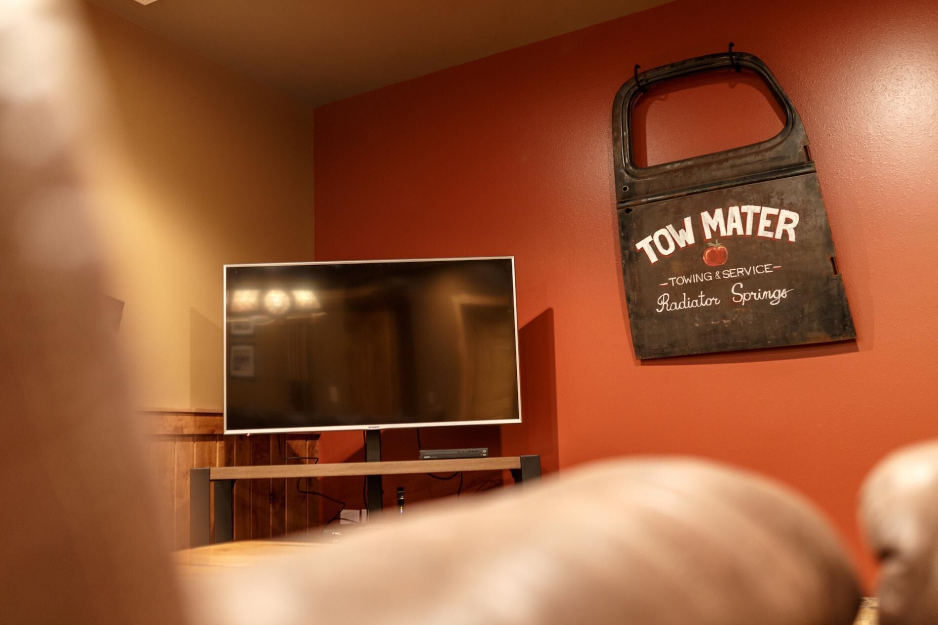 A room with a flat-screen TV mounted on a wooden stand, a wall-mounted sign that reads "TOW MATER TOWING & SERVICE Radiator Springs", and a partial view of a dog in the foreground.