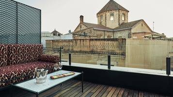 Una terraza en la azotea con un sofá estampado, una mesa con una copa de vino y una bandeja de frutas, y vistas a un edificio histórico con una torre.