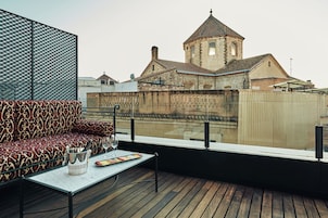 Une terrasse sur le toit, avec un canapé à motifs, une table sur laquelle se trouvent un verre à vin et une assiette de fruits, et une vue sur un bâtiment historique doté d’une tour.