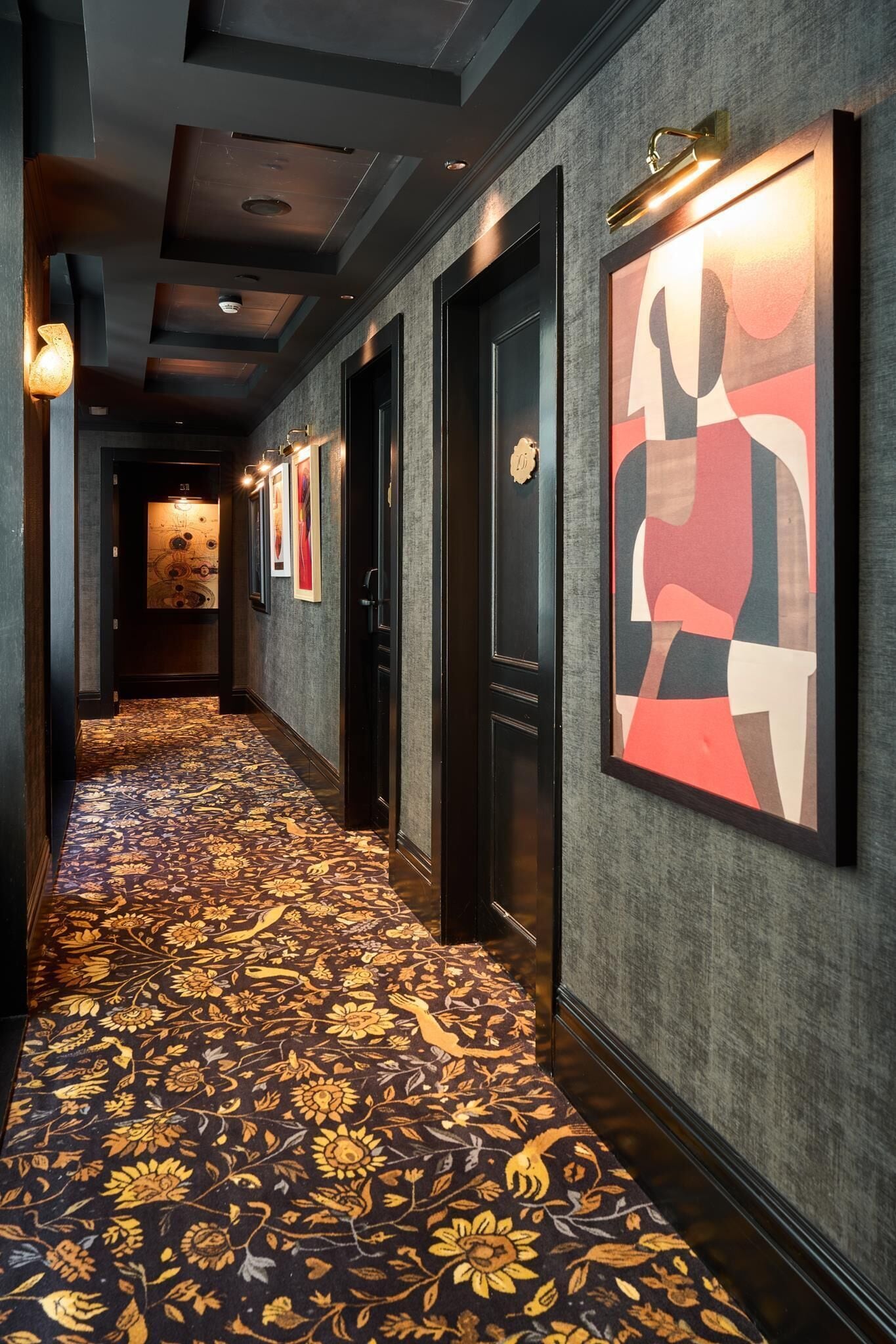 A hotel corridor with a patterned carpet, framed artwork on the walls, and a door with a number plaque.