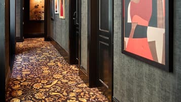 A hotel corridor with a patterned carpet, framed artwork on the walls, and a door with a number plaque.