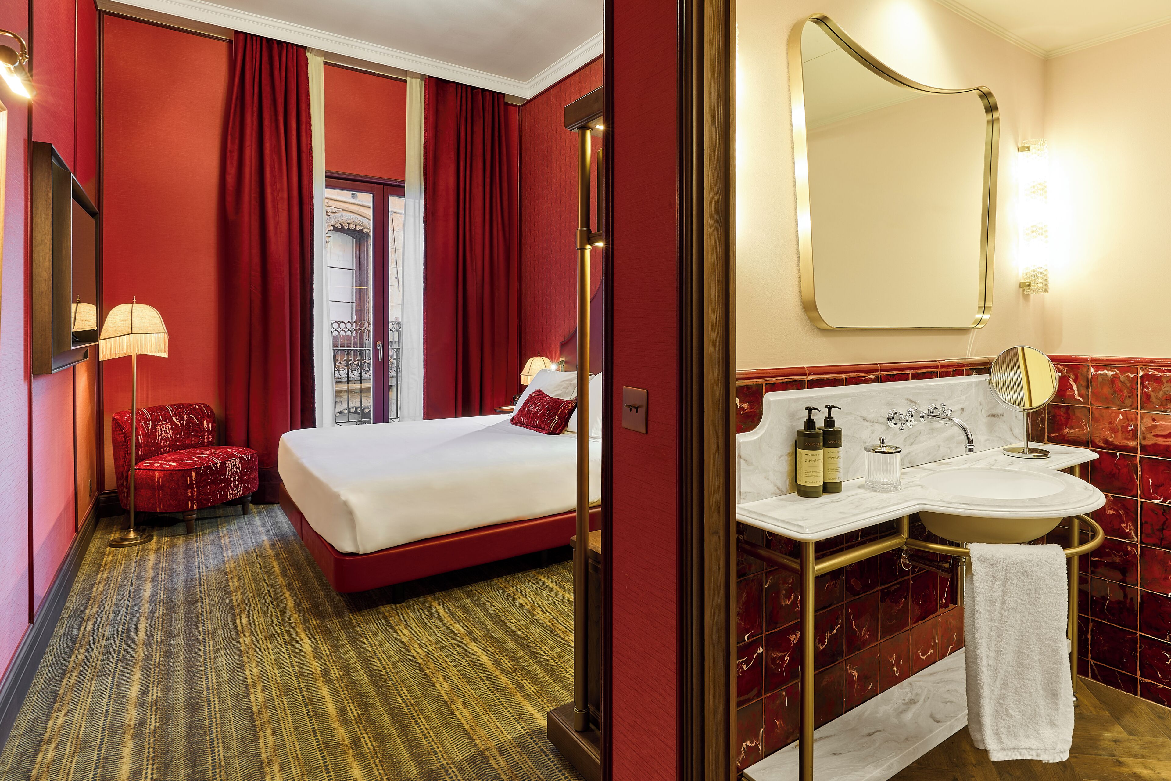 A hotel room with a bed, a red chair, a bathroom with a sink and mirror, and a view of a balcony.