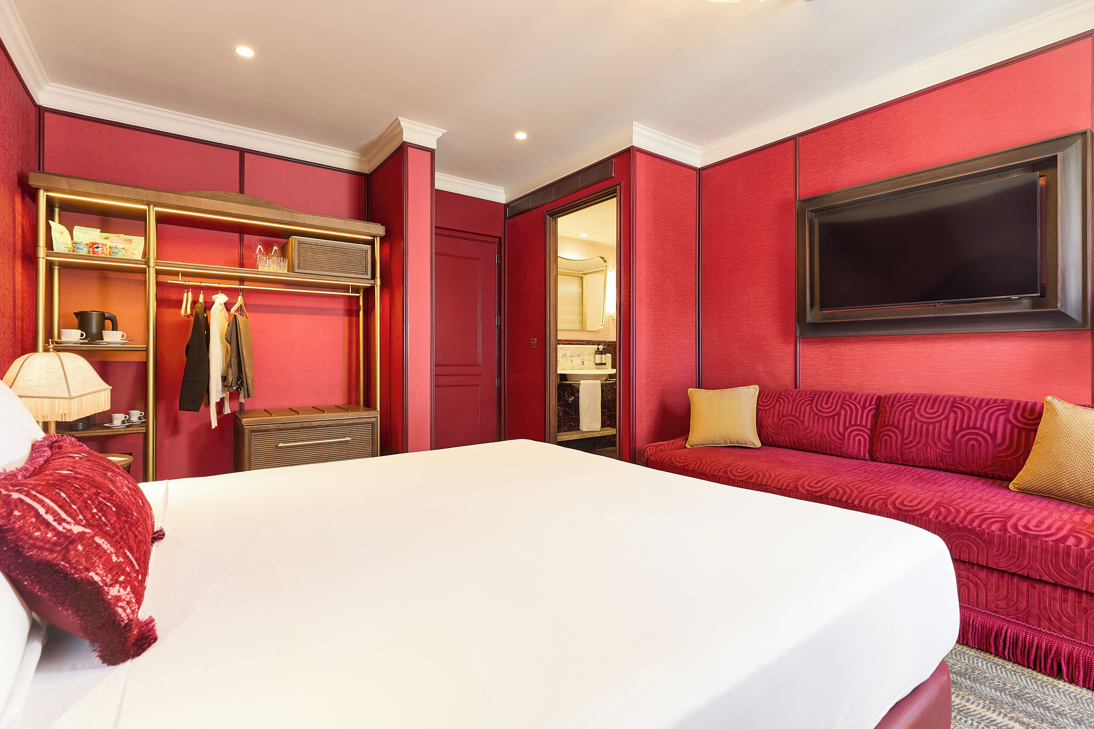A hotel room with a large bed, a red sofa, a TV mounted on the wall, and a wardrobe with shelves.