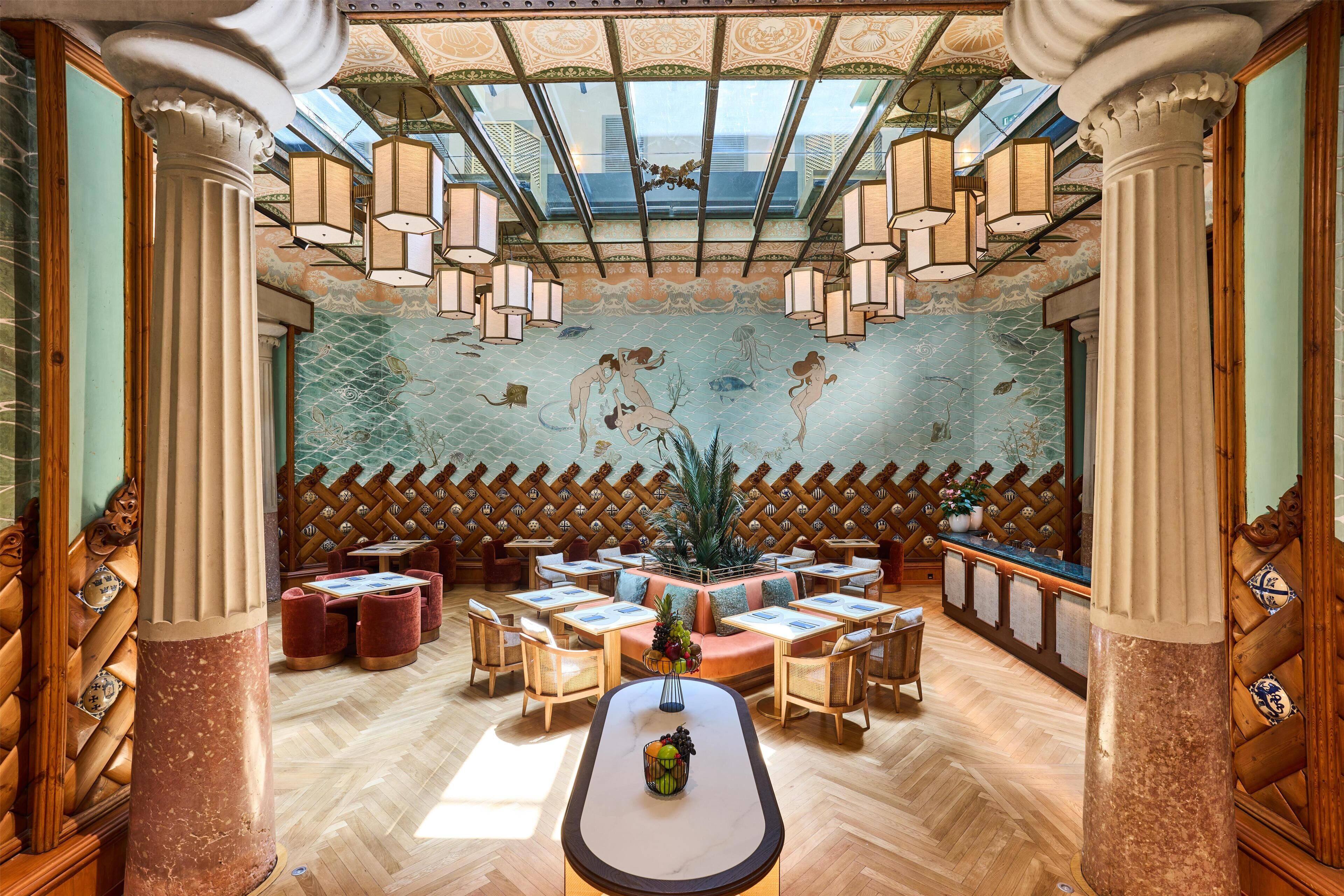 Een restaurant met een hoge, versierde plafond en een muurschildering van zeedieren. Tafels staan rondom een centraal eiland met een fruitschaal.