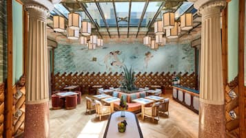 Een restaurant met een hoge, versierde plafond en een muurschildering van zeedieren. Tafels staan rondom een centraal eiland met een fruitschaal.